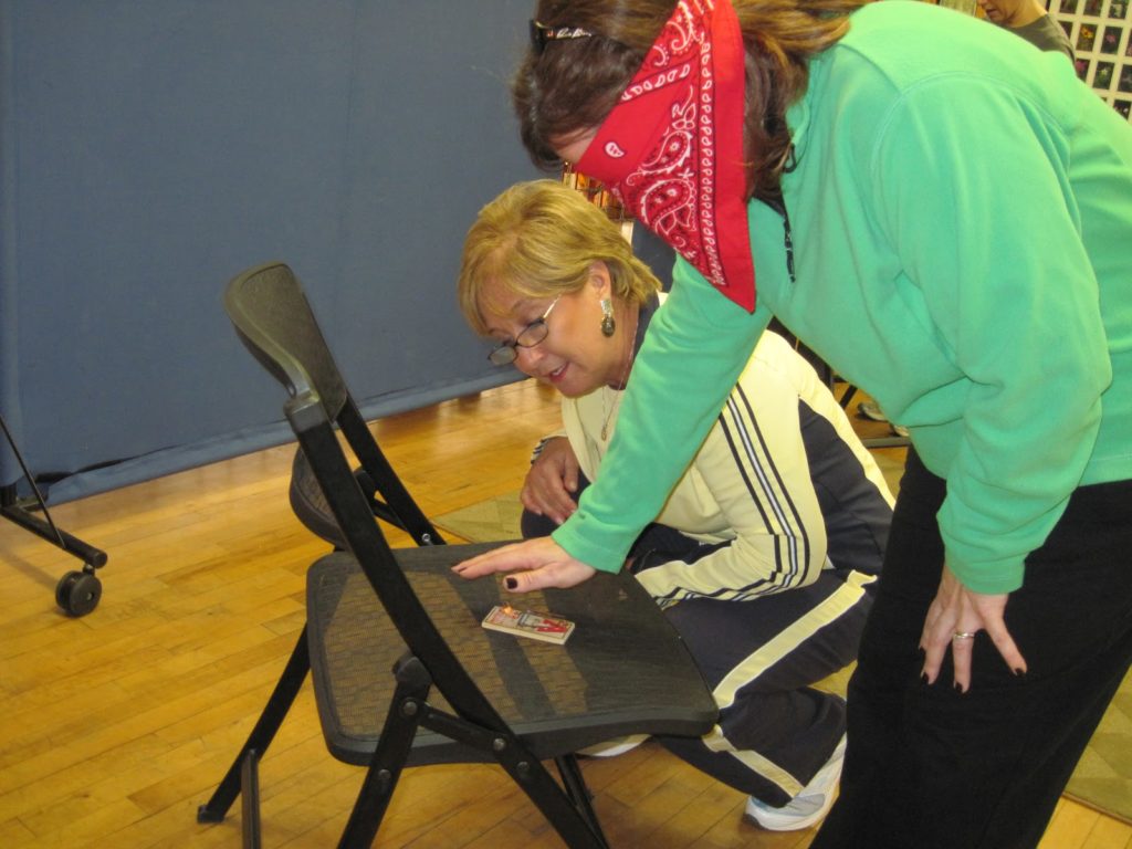 One woman instructing a blindfolded woman how to deactivate a mousetrap.