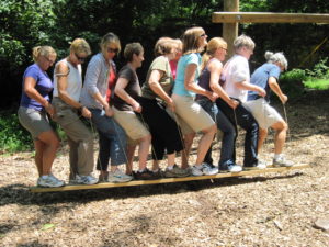 Team members stand single file with one foot on each board, walking as one unit.