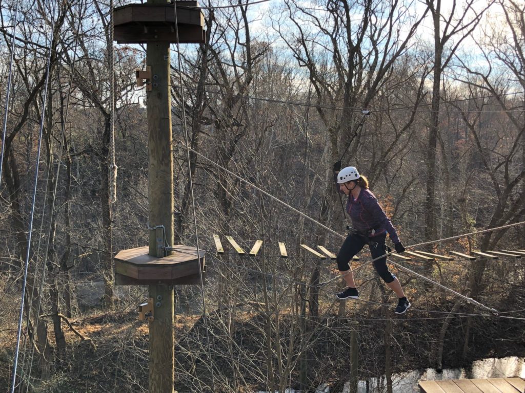 A woman high up on a ropes challenge course, suspended on a single wire.