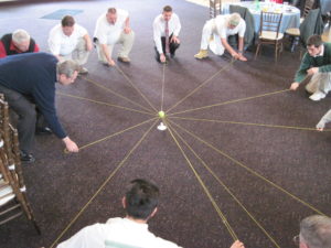 A group of men in business clothes try to suspend a ball on a ring, supported by outstretched pieces of string.