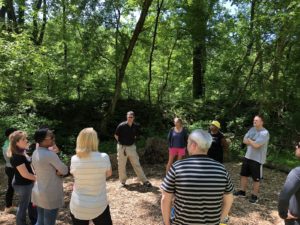 A group of people standing in a circle outdoors, surrounded by trees, facing the group facilitator.